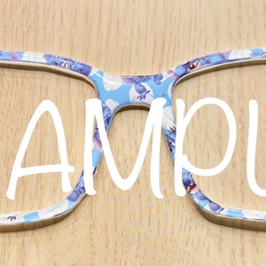 Puede incluir: Montura de gafas con un patr&oacute;n azul claro de peque&ntilde;os personajes de dibujos animados y la palabra SAMPLE impresa en letras blancas grandes en el centro. La montura tiene forma rectangular con esquinas redondeadas y acabado liso.