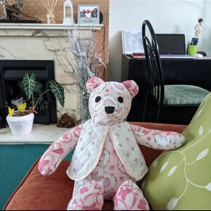 May include: A pink and white patchwork teddy bear wearing a white quilted waistcoat with a floral pattern. The bear is sitting on a pink sofa with a green cushion.