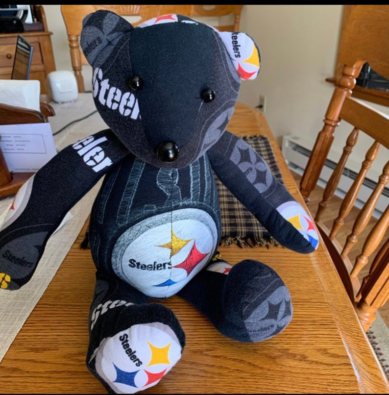 May include: A black teddy bear made from Steelers fabric. The bear has black button eyes and a black nose. The bear is sitting on a wooden table.