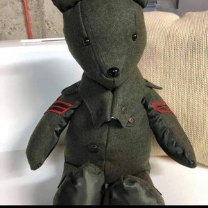 May include: A plush green teddy bear wearing a green jacket with red trim and black buttons. The bear has black eyes and a black nose.