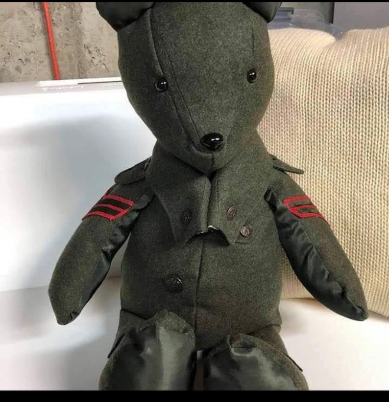May include: A plush green teddy bear wearing a green jacket with red trim and black buttons. The bear has black eyes and a black nose.