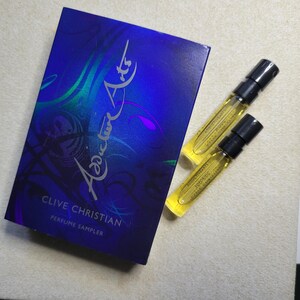 May include: A blue box with the text "Clive Christian Perfume Sampler" and the word "Addictive" written in a stylized font. Two small glass perfume sample bottles with black caps are in front of the box.