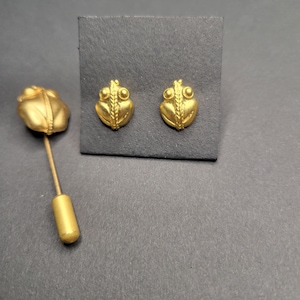 May include: A set of gold-toned jewelry featuring two frog-shaped stud earrings and one matching frog-shaped lapel pin. Each piece displays a textured, matte finish with a central vertical ridge and two prominent circular eyes on the frog head.
