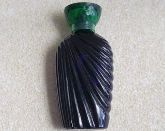 May include: A small, dark blue glass perfume bottle with a distinct diagonal ribbed texture across its body. The bottle is topped with a translucent, faceted emerald green glass cap that features a honeycomb pattern on its top surface.