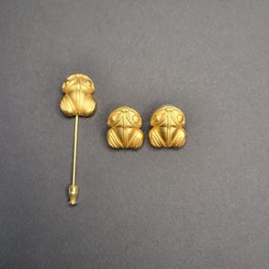 May include: A set of gold-toned jewelry consisting of one stick pin and two stud earrings, each featuring a stylized frog design with a smooth, matte finish and symmetrical embossed lines on the surface of the frog-shaped metal components.