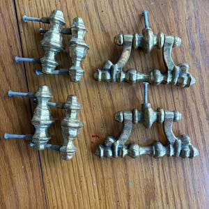 May include: Four antique brass drawer pulls with ornate detailing. Two pulls have a vertical design with screws, and two have a horizontal design with screws. The brass has a slightly aged appearance.