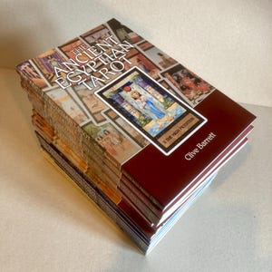 May include: A stack of paperback books titled The Ancient Egyptian Tarot by Clive Barrett. The cover features a collage of tarot card illustrations and a central image of The High Priestess card. The books have a deep red spine and cover section.