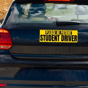 May include: A black car's rear with a yellow and black sign that reads "PLEASE BE PATIENT STUDENT DRIVER". The sign is rectangular and has a black border. The car's taillights are red.