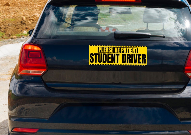 May include: A black car's rear with a yellow and black sign that reads "PLEASE BE PATIENT STUDENT DRIVER". The sign is rectangular and has a black border. The car's taillights are red.
