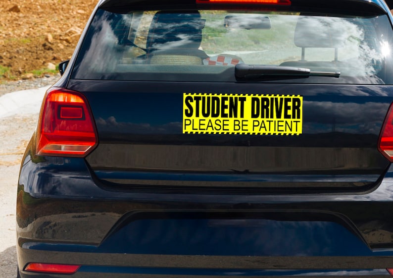 May include: A black car's rear with a yellow and black rectangular sticker. The sticker reads "STUDENT DRIVER PLEASE BE PATIENT" in bold, black capital letters. The car's taillights are red.