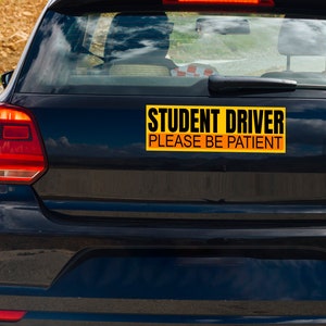 May include: A black car with a yellow and black bumper sticker that reads "STUDENT DRIVER PLEASE BE PATIENT". The car's taillights are red, and the background shows a road and sky.