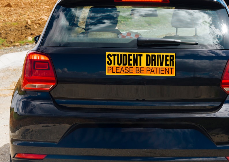 May include: A black car with a yellow and black bumper sticker that reads "STUDENT DRIVER PLEASE BE PATIENT". The car's taillights are red, and the background shows a road and sky.