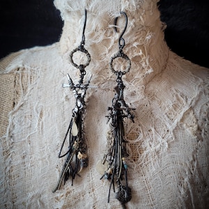 May include: A pair of handcrafted, dark metal earrings with intricate designs. Each earring features a circular top, a cross-like structure, and dangling charms, beads, and metal accents. The earrings are displayed on a textured, off-white surface.
