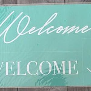 Puede incluir: Un letrero rectangular turquesa con la palabra "Welcome" en escritura blanca y letras de bloque. La palabra "home" está escrita verticalmente en escritura en el lado derecho del letrero. El letrero está en una funda de plástico transparente.