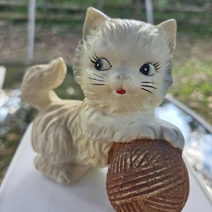 May include: A cream-colored ceramic cat figurine with blue eyes, long eyelashes, and a small red mouth. The cat is standing on all fours with its tail raised and one front paw resting on a textured, round, brown ball of yarn with carved details.