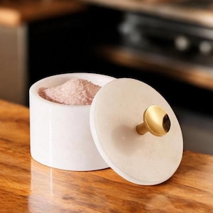 Könnte beinhalten: Ein weißer Salzbehälter aus Marmor mit Deckel. Der Deckel hat einen goldfarbenen Knopf. Der Behälter ist offen und zeigt rosa Salz. Der Salzbehälter steht auf einer Holzoberfläche.