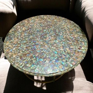 Könnte beinhalten: Runder Beistelltisch mit schimmernder, irisierender Oberfläche. Die Tischplatte besteht aus kleinen, farbigen Teilen, die einen Mosaikeffekt erzeugen. Das Gestell ist aus goldfarbenem Metall. Ein dekoratives Möbelstück.