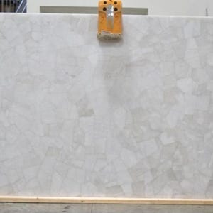 May include: A large, rectangular slab of light grey stone with a mosaic pattern. The stone is held by an orange lifting device. The surface has a textured appearance, with irregular shapes and colour variations. The slab is likely 200 cm x 100 cm.