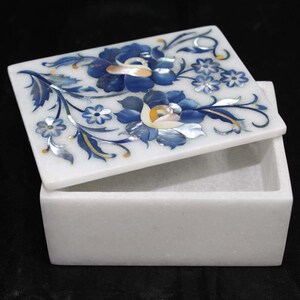 Könnte beinhalten: Eine weiße Marmorbox mit einem Klappdeckel, verziert mit einem floralen Design. Der Deckel zeigt blaue Blumen mit Perlmuttakzenten und blaue Blätter. Die rechteckige Box ist geöffnet und zeigt ihr Inneres.