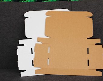 May include: Two flat-pack cardboard boxes, one white and one brown, are shown unfolded on a dark grey surface. The boxes have flaps and slots for easy assembly.