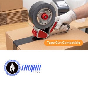 Op de afbeelding: Een handmatige tape-dispenser met een rol zwarte tape, voorzien van een rood en grijs kunststof frame. Het apparaat brengt tape aan op een bruine kartonnen doos. Een oranje label op de doos vermeldt Tape Gun Compatible. Het Trojan Packaging logo is zichtbaar.