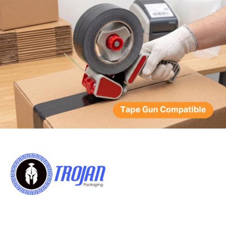 Op de afbeelding: Een handmatige tape-dispenser met een rol zwarte tape, voorzien van een rood en grijs kunststof frame. Het apparaat brengt tape aan op een bruine kartonnen doos. Een oranje label op de doos vermeldt Tape Gun Compatible. Het Trojan Packaging logo is zichtbaar.