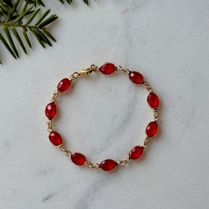 May include: A gold bracelet with oval red gemstones. The bracelet is linked with small gold connectors and has a clasp. The jewellery is displayed on a white marble surface with a green leafy branch in the background.