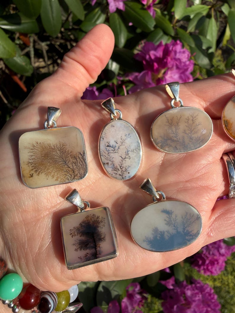 May include: A collection of silver pendants with dendritic agate stones. The pendants have various shapes, including square and oval, and feature unique, tree-like patterns in shades of brown, black, and white. The pendants are displayed against a background of green foliage and purple flowers.
