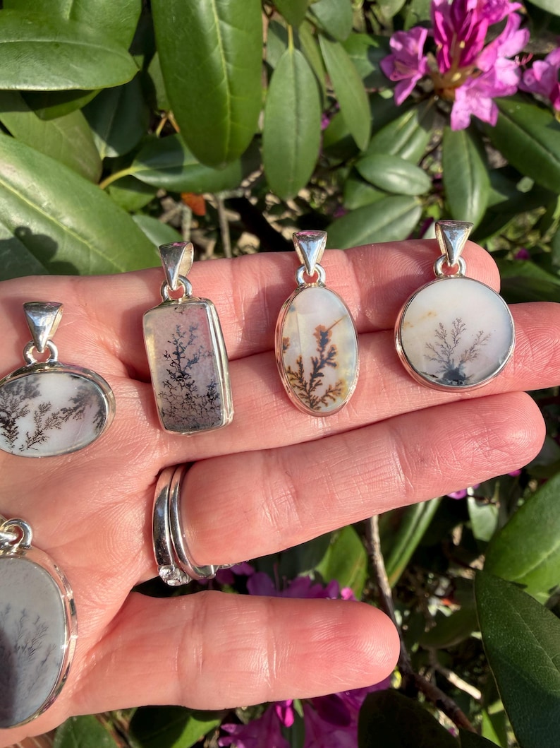 May include: A collection of silver pendants with dendritic agate stones. The pendants have various shapes, including oval, rectangular, and round, each showcasing unique, tree-like patterns in shades of black, white, and brown. The pendants are displayed against a backdrop of green foliage and pink flowers.
