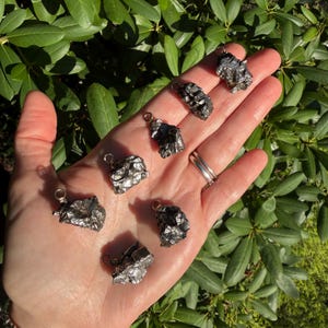 May include: Seven small, irregular, metallic-gray meteorite pendants, each featuring a silver-toned metal bail attached to the top. The pieces have a rough, textured surface with a dark, reflective finish, showcasing unique, jagged, and natural shapes.
