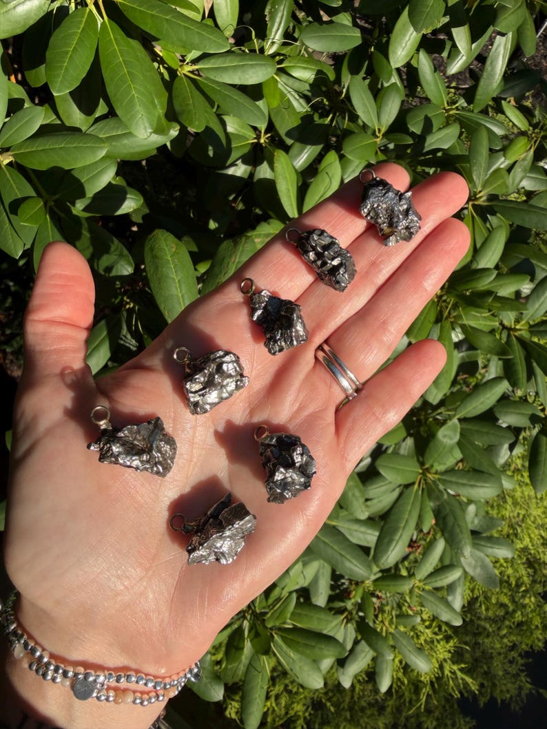 May include: Seven small, irregular, metallic-gray meteorite pendants, each featuring a silver-toned metal bail attached to the top. The pieces have a rough, textured surface with a dark, reflective finish, showcasing unique, jagged, and natural shapes.