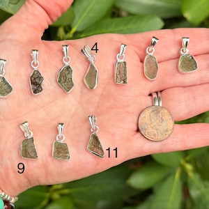 May include: A collection of eleven small, irregular, olive-green moldavite pendants set in silver bails, numbered 1, 4, 8, 9, and 11. A copper penny is included for scale, showing the relative size of these textured, translucent gemstone pieces.