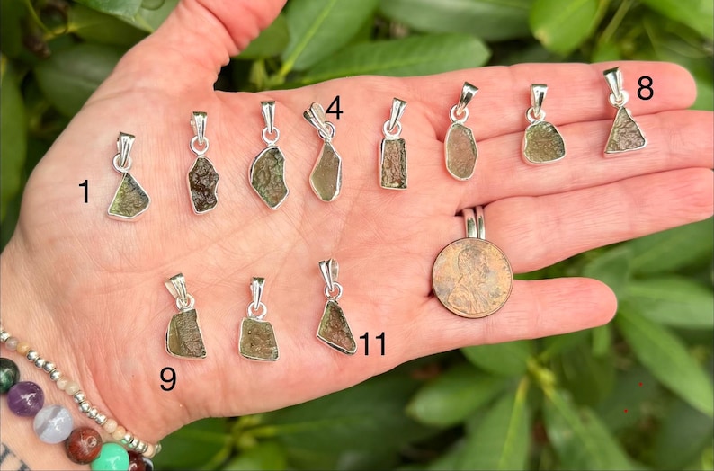 May include: A collection of eleven small, irregular, olive-green moldavite pendants set in silver bails, numbered 1, 4, 8, 9, and 11. A copper penny is included for scale, showing the relative size of these textured, translucent gemstone pieces.