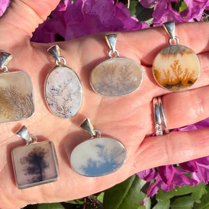 May include: A collection of dendritic agate pendants with silver settings. The pendants feature unique, tree-like patterns in shades of brown, white, and gray against a translucent background. The pendants are displayed in a hand, with a backdrop of pink flowers.