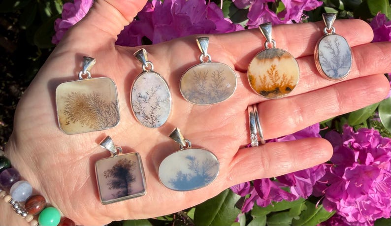 May include: A collection of dendritic agate pendants with silver settings. The pendants feature unique, tree-like patterns in shades of brown, white, and gray against a translucent background. The pendants are displayed in a hand, with a backdrop of pink flowers.