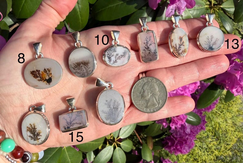 May include: A collection of dendritic agate pendants with silver settings, showcasing unique patterns resembling miniature landscapes. The pendants vary in shape, including oval, rectangular, and round designs. A US quarter is included for size comparison. The pendants are displayed against a hand.