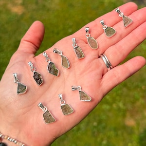 May include: A collection of eleven raw, olive-green moldavite pendants, each set in a simple silver-toned metal bail. The stones feature irregular, textured surfaces and varied shapes, ranging from deep forest green to lighter translucent olive tones.