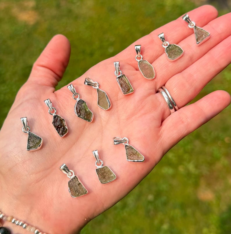 May include: A collection of eleven raw, olive-green moldavite pendants, each set in a simple silver-toned metal bail. The stones feature irregular, textured surfaces and varied shapes, ranging from deep forest green to lighter translucent olive tones.