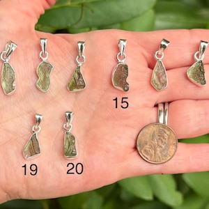 May include: A collection of nine raw, green, translucent stone pendants set in silver-toned metal bails, labelled with numbers 12, 15, 18, 19, and 20. A copper-coloured coin is included for scale, showing the relative size of the small jewellery pieces.