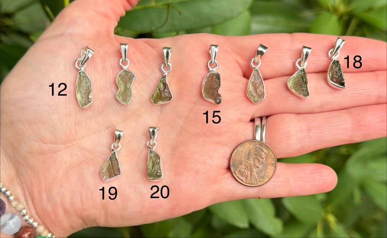 May include: A collection of nine raw, green, translucent stone pendants set in silver-toned metal bails, labelled with numbers 12, 15, 18, 19, and 20. A copper-coloured coin is included for scale, showing the relative size of the small jewellery pieces.