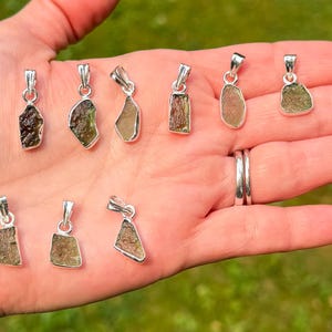 May include: A collection of eleven small, raw, olive-green stone pendants, each set in a simple silver-toned metal bail. The stones vary in shape and texture, displaying natural, irregular edges and earthy, translucent green tones across the set.