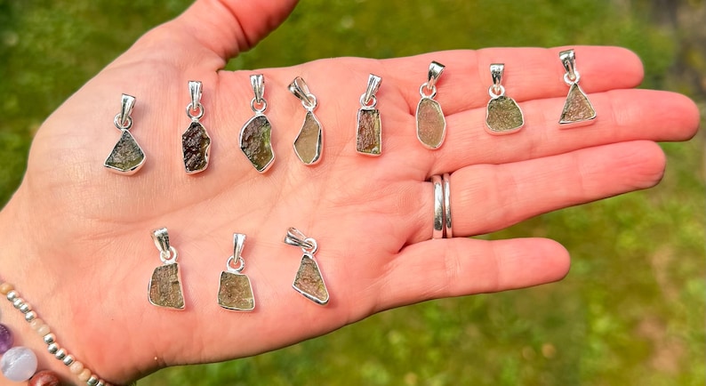 May include: A collection of eleven small, raw, olive-green stone pendants, each set in a simple silver-toned metal bail. The stones vary in shape and texture, displaying natural, irregular edges and earthy, translucent green tones across the set.