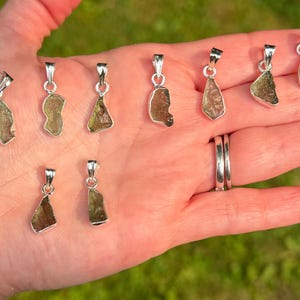 May include: A collection of nine small, irregular, translucent olive-green moldavite pendants, each set in a polished silver-toned metal bail. The pendants vary in shape and texture, displaying natural, rough-hewn surfaces held in a palm.