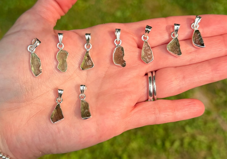 May include: A collection of nine small, irregular, translucent olive-green moldavite pendants, each set in a polished silver-toned metal bail. The pendants vary in shape and texture, displaying natural, rough-hewn surfaces held in a palm.