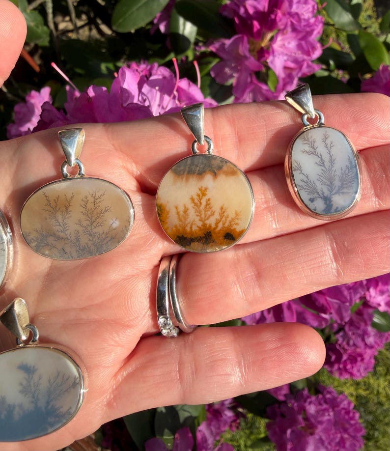 May include: Silver pendants with dendritic patterns. The pendants have oval and round shapes, showcasing natural, tree-like inclusions in shades of beige, brown, and gray. The pendants are displayed against a backdrop of vibrant pink flowers.