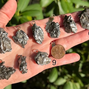 May include: A collection of nine irregular, metallic-toned meteorite pendants, each featuring a small circular bail for suspension. The pieces are numbered one through nine, with a copper penny placed next to number nine for scale reference.