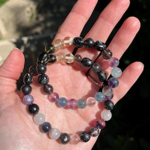 May include: Two beaded bracelets with a variety of colorful gemstones. The bracelets feature a mix of black, purple, clear, and other colored beads. The bracelets are held in a hand.