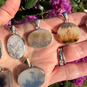May include: A collection of dendritic agate pendants with silver-colored settings. The pendants feature unique, tree-like patterns in shades of brown, white, and gray. The pendants are oval, round, and square shaped. The background includes green leaves and pink flowers.