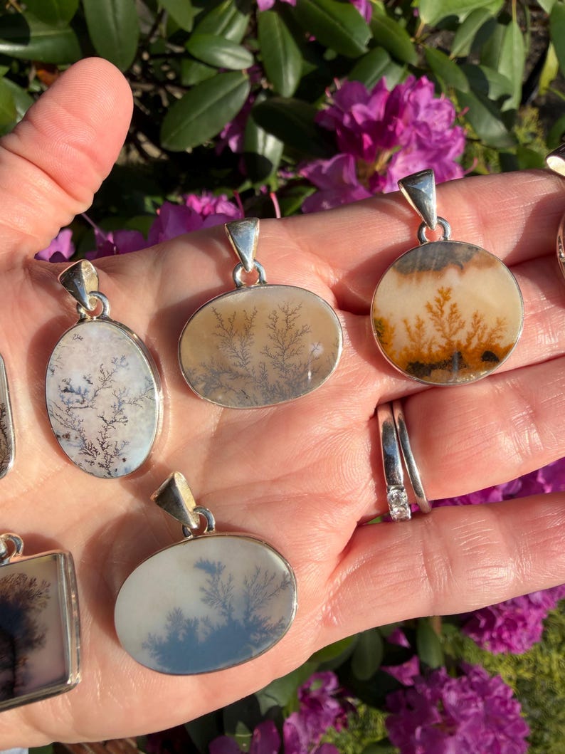 May include: A collection of dendritic agate pendants with silver-colored settings. The pendants feature unique, tree-like patterns in shades of brown, white, and gray. The pendants are oval, round, and square shaped. The background includes green leaves and pink flowers.
