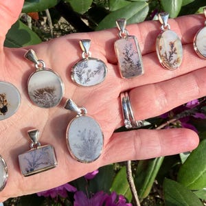 May include: A collection of silver pendants with dendritic agate stones. The pendants have various shapes, including oval, rectangular, and circular. The agate stones display unique, fern-like patterns in shades of brown, gray, and white. The pendants are displayed on an open hand.